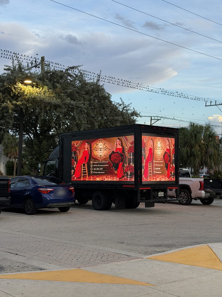 Black box truck with lit LED screens on the rear and passenger side, parked at dusk with cars alongside